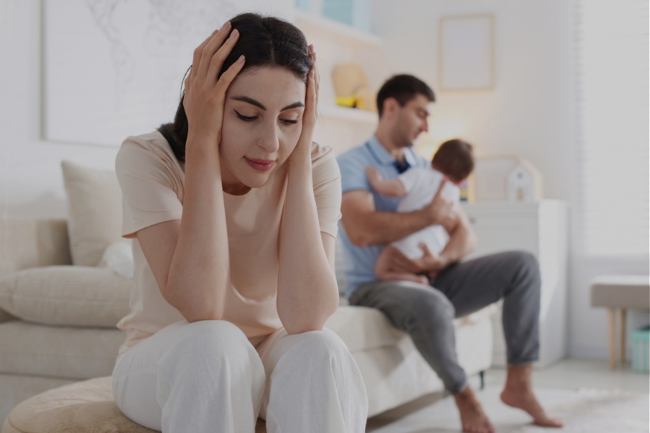 Overwhelmed mother experiencing mental overload while partner holds baby in background, illustrating the default parent dynamic