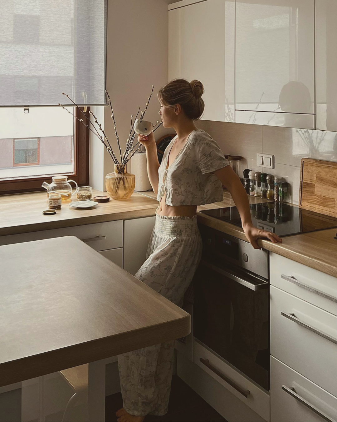 Young woman in pajamas drinking coffee in a cozy kitchen at sunrise, leaning on the counter and slowly starting her morning.