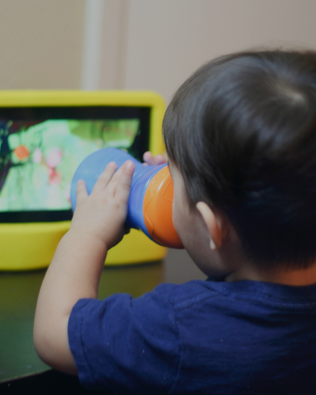 Toddler drinking from a sippy cup while watching a kids’ show on a tablet screen, morning screen time for an exhausted parent.