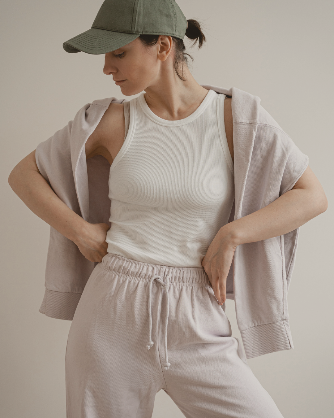 Tired mom in soft neutral loungewear and a baseball cap, standing with hands on hips in a relaxed but functional comfy outfit.