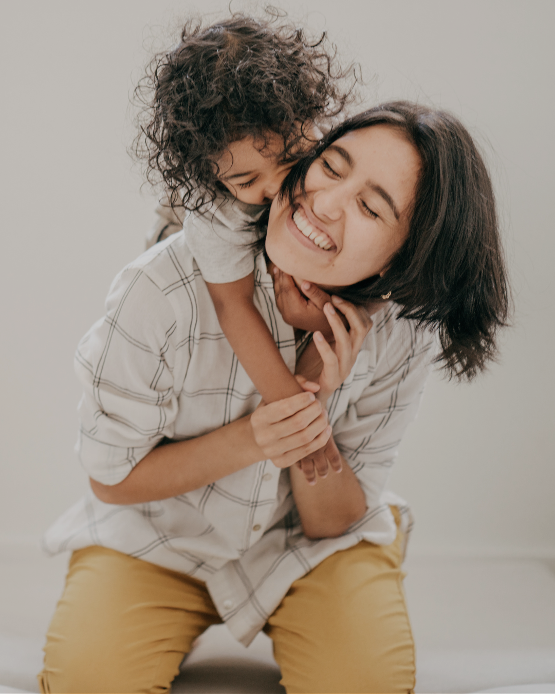 Smiling mother holding young child with curly hair in warm embrace, both with eyes closed, wearing casual clothing in neutral tones against soft gray background