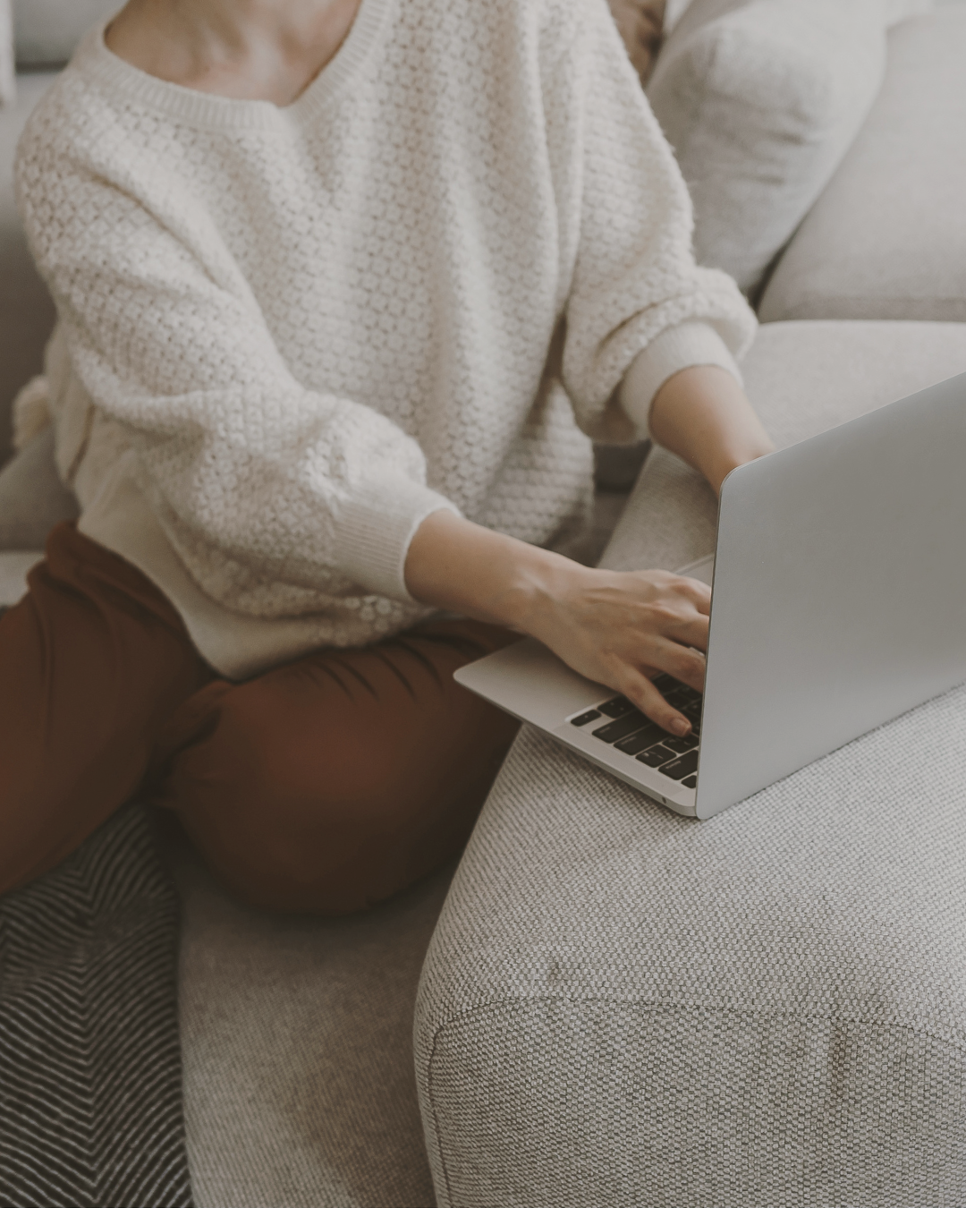 Person wearing cream knit sweater working on laptop while sitting on beige couch, view from torso showing hands on keyboard in cozy home environment