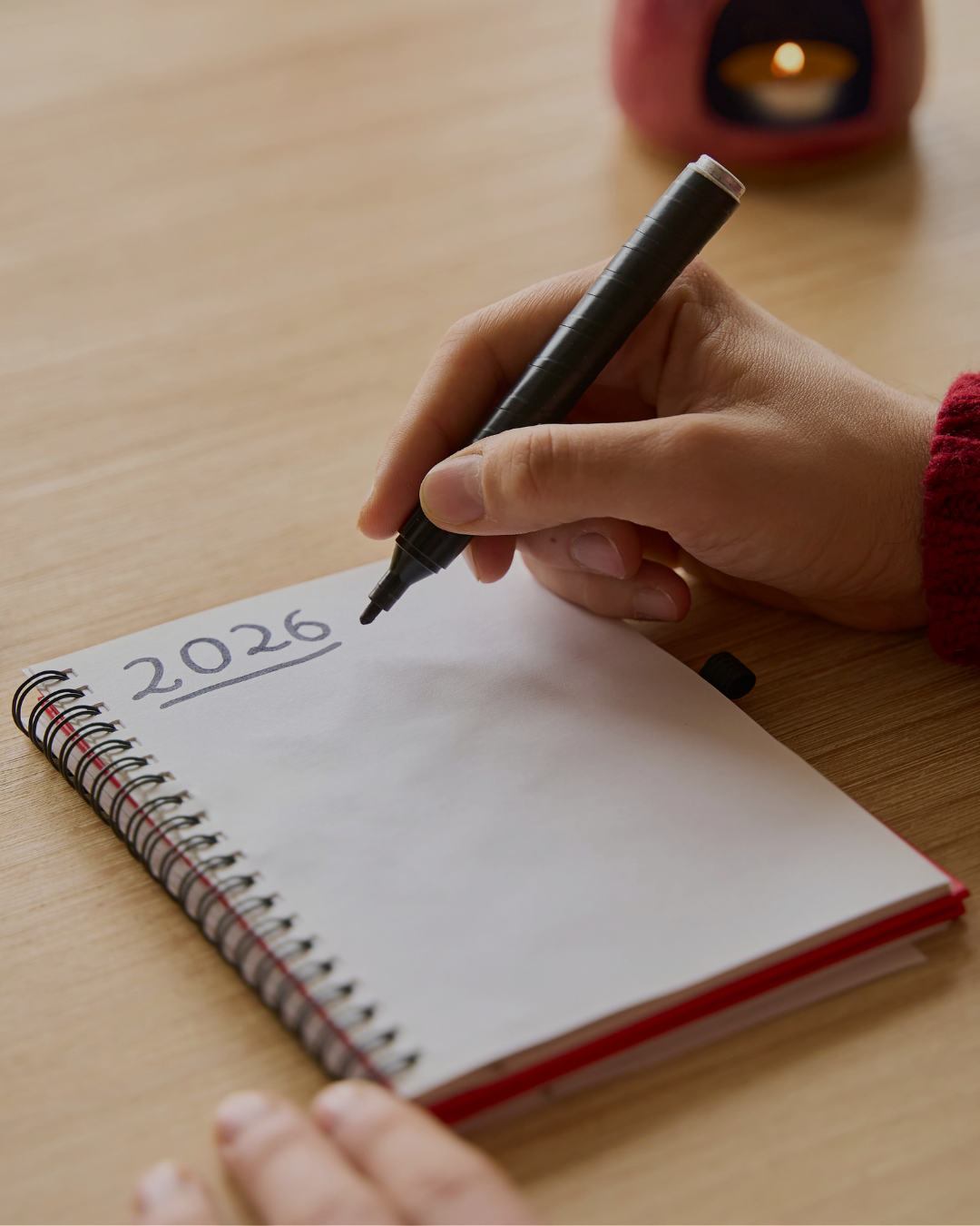 Close-up of hand writing "2026" with black marker in spiral notebook, candle softly blurred in background, planning and goal-setting concept