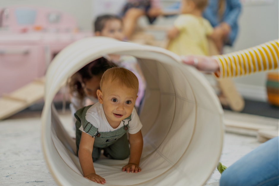 Baby in white and green outfit crawling through a white play tunnel while caregiver watches in background, representing executive function challenges for AUDHD moms
