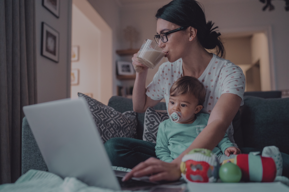 Tired mom drinking coffee while holding toddler on her lap and working on laptop at home, with toys scattered on couch