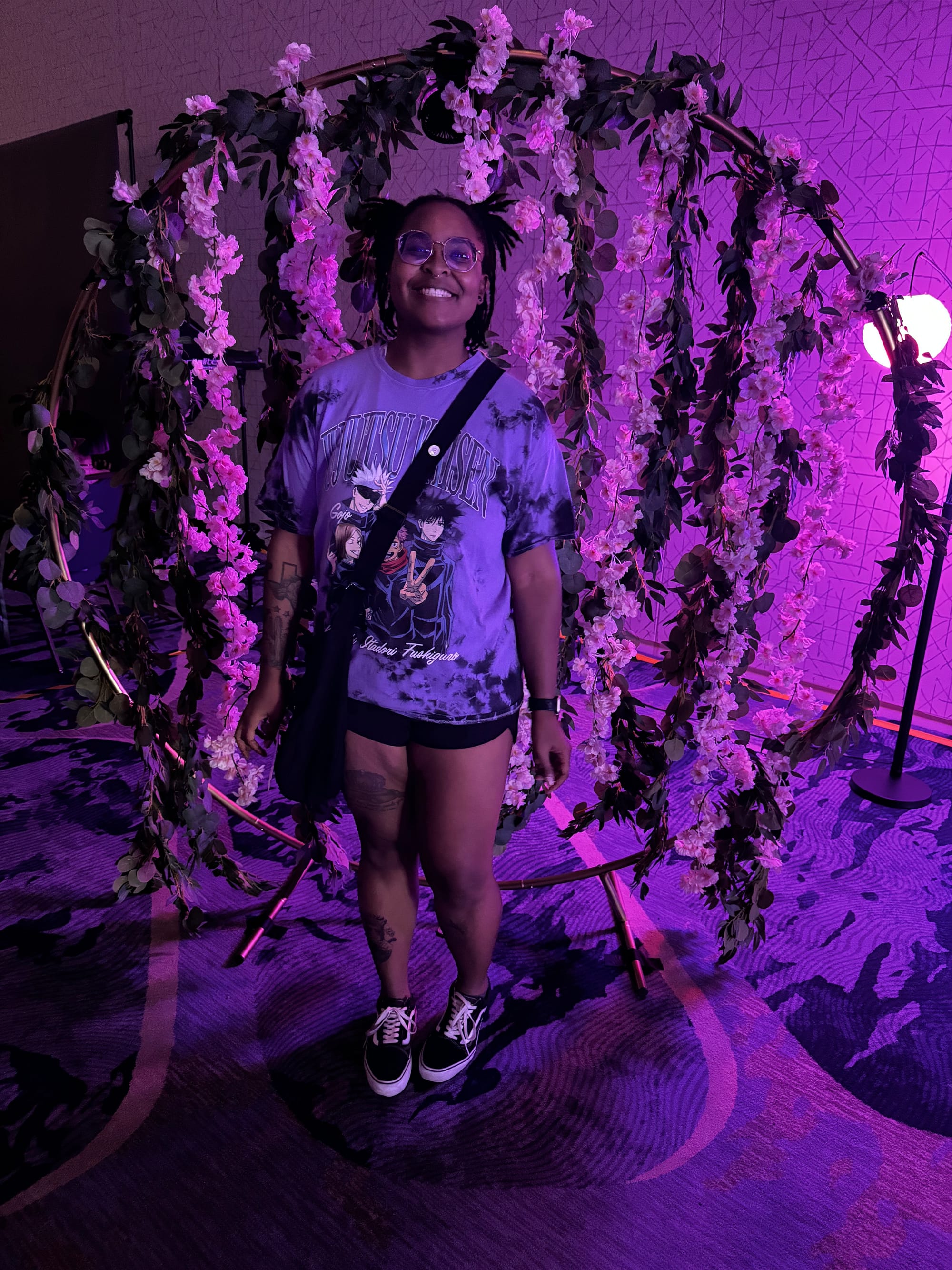 Shae standing in front of purple flower backdrop wearing a Jujutsu Kaisen anime t-shirt, glasses, and showing visible tattoos