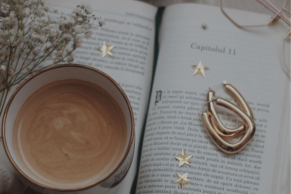 Overhead view of open book with gold jewelry, coffee cup, and dried flowers arranged on neutral surface, peaceful reading moment aesthetic