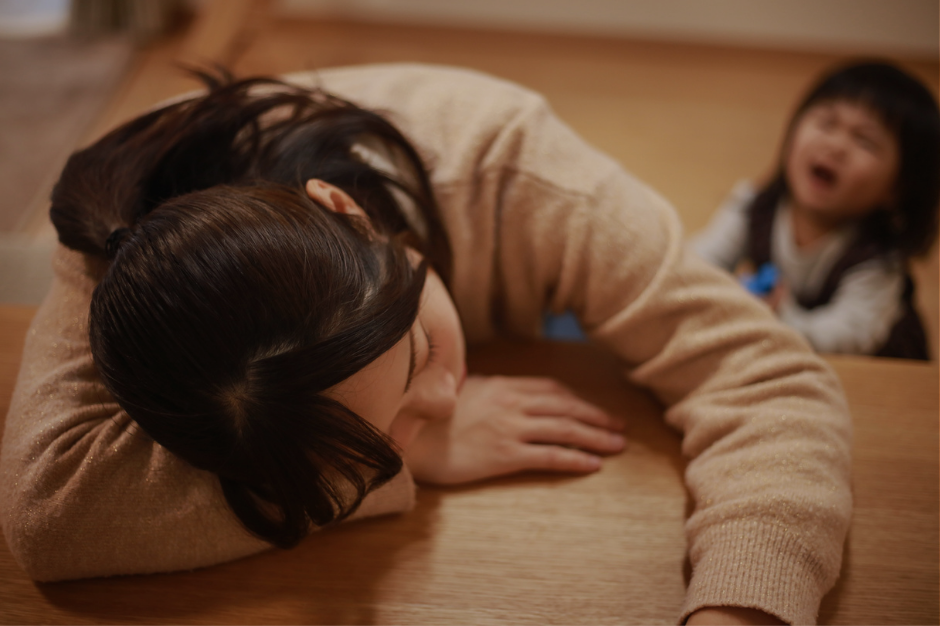 Exhausted mother lying face down on hardwood floor with young child in background - overwhelmed mom experiencing sensory overload and feeling touched out