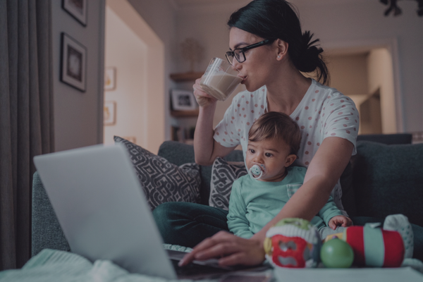 Tired mom drinking coffee while holding toddler on her lap and working on laptop at home, with toys scattered on couch
