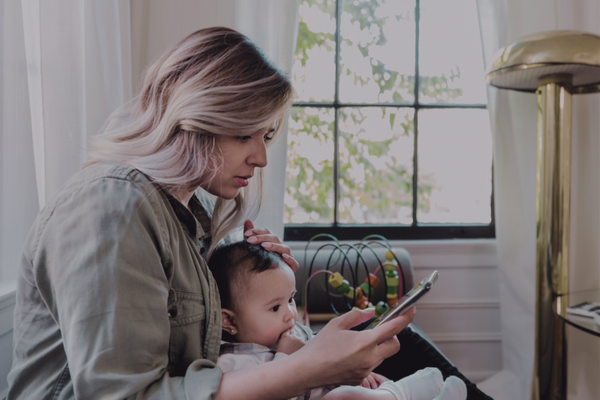 Mom with highlighted hair holding baby while looking at smartphone, sitting near window with natural light