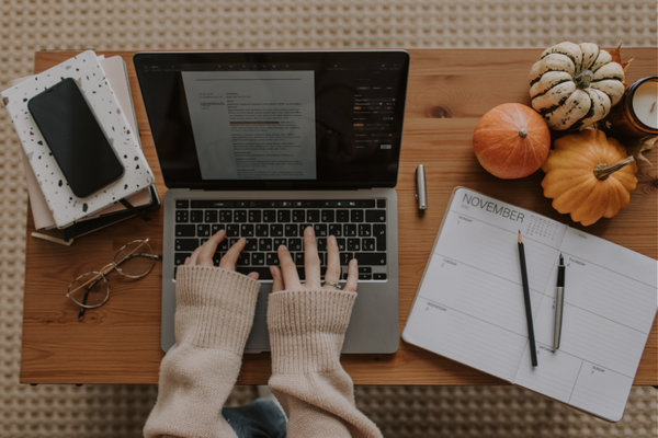 Hands typing on a laptop at a desk with an open planner to the right and a stack of books and a phone on top to the left of the hands