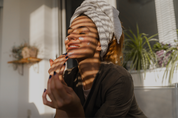 Woman in gray towel wrap and dark loungewear relaxing by window with soft natural light and plants, representing post-holiday rest and recovery for neurodivergent moms