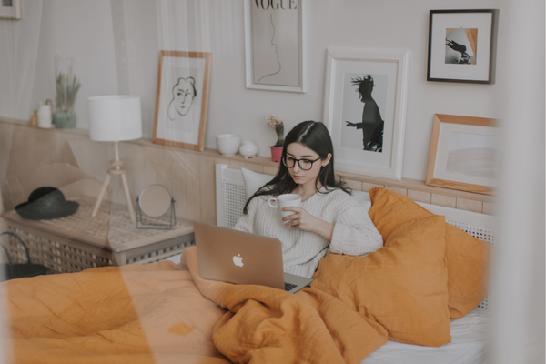 Neurodivergent woman with glasses sits in bed with laptop and coffee, representing executive dysfunction and working from home in comfortable environment with yellow bedding