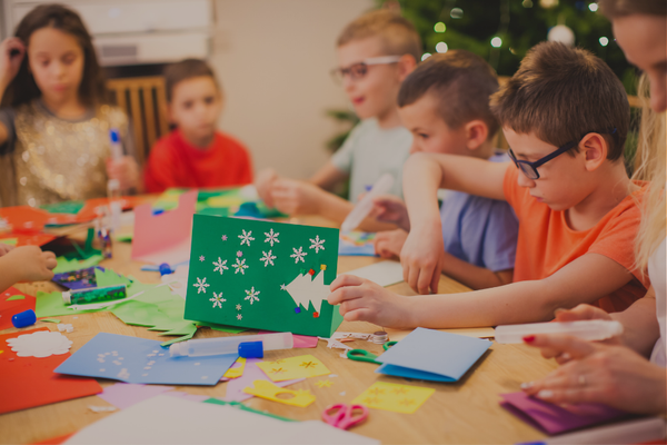 Group of children sitting at a table doing arts and crafts for the Christmas holiday season with a Christmas tree in the back